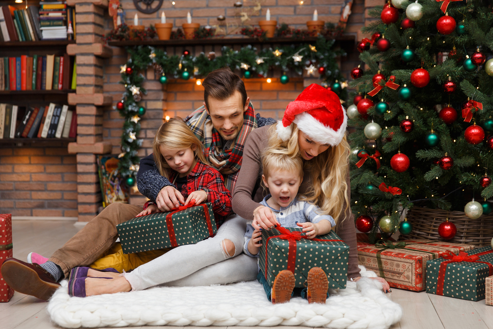 Mom, dad, and kids sit by Christmas tree and open presents