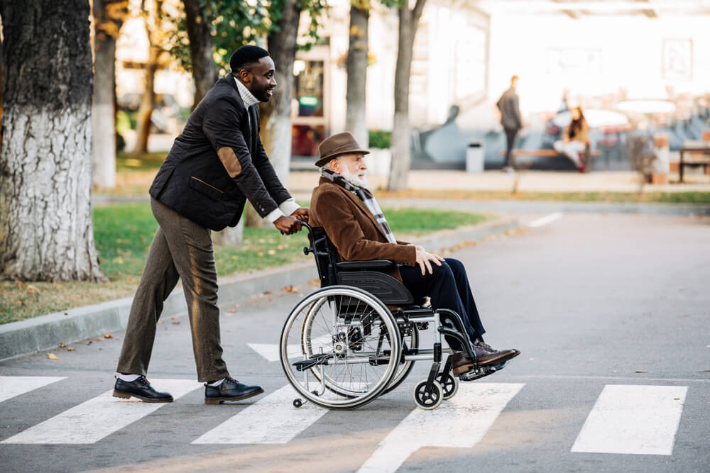 man helps elderly man in wheelchair across street