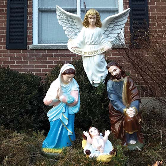 An outdoor Nativity set displayed in front of a brick home: Mary kneeling in prayer and Joseph seated beside the Baby Jesus in the manger, with an angel above them.